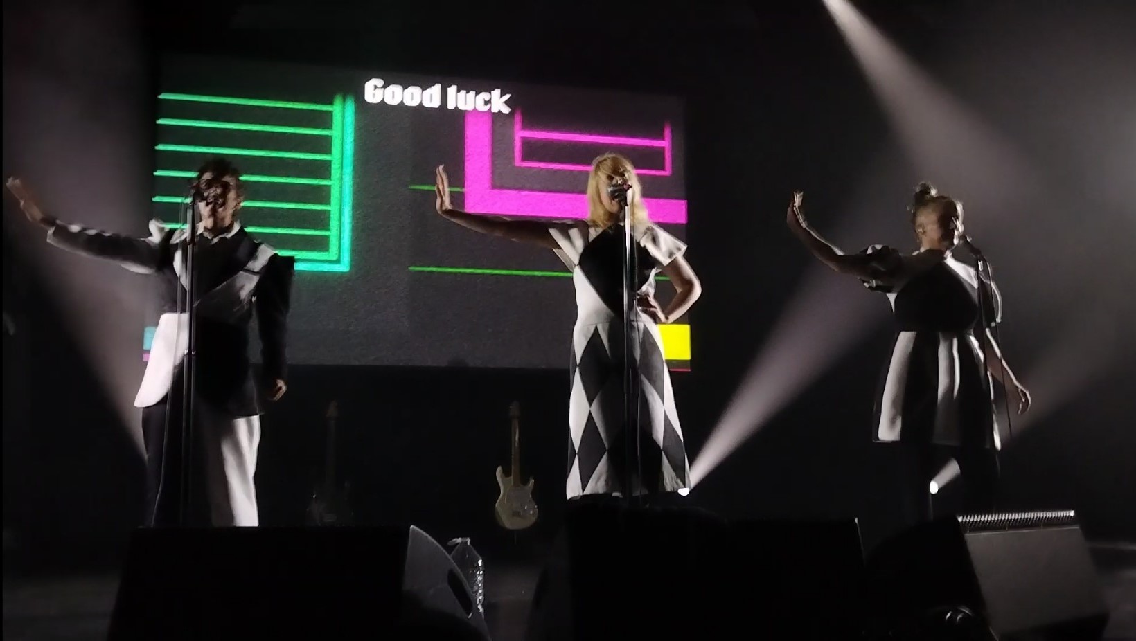 Photo of a Le Tigre concert. JD Samson, Johanna Fateman, and Kathleen Hanna are performing in front of a lyrics screen which reads Good luck.