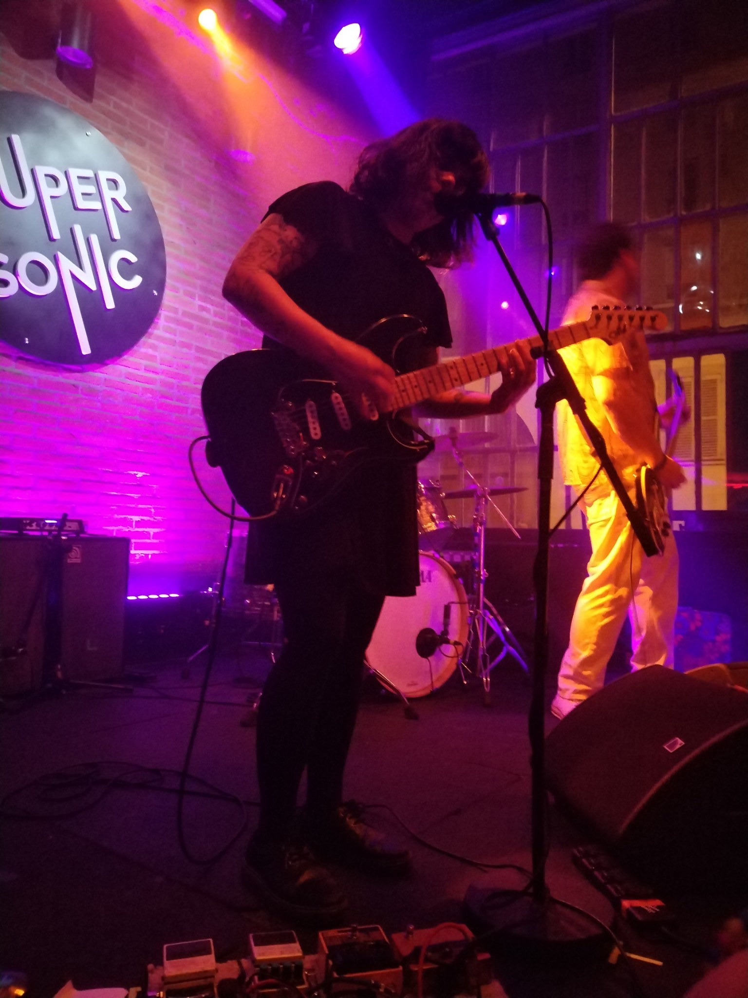 Photo of Marissa Paternoster playing guitar and singing in a bar, the lighting is very pink and purple